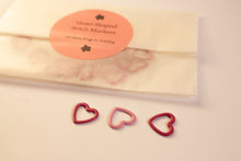 Load image into Gallery viewer, Heart-shaped pink metal stitch markers with a paper envelope on a light background
